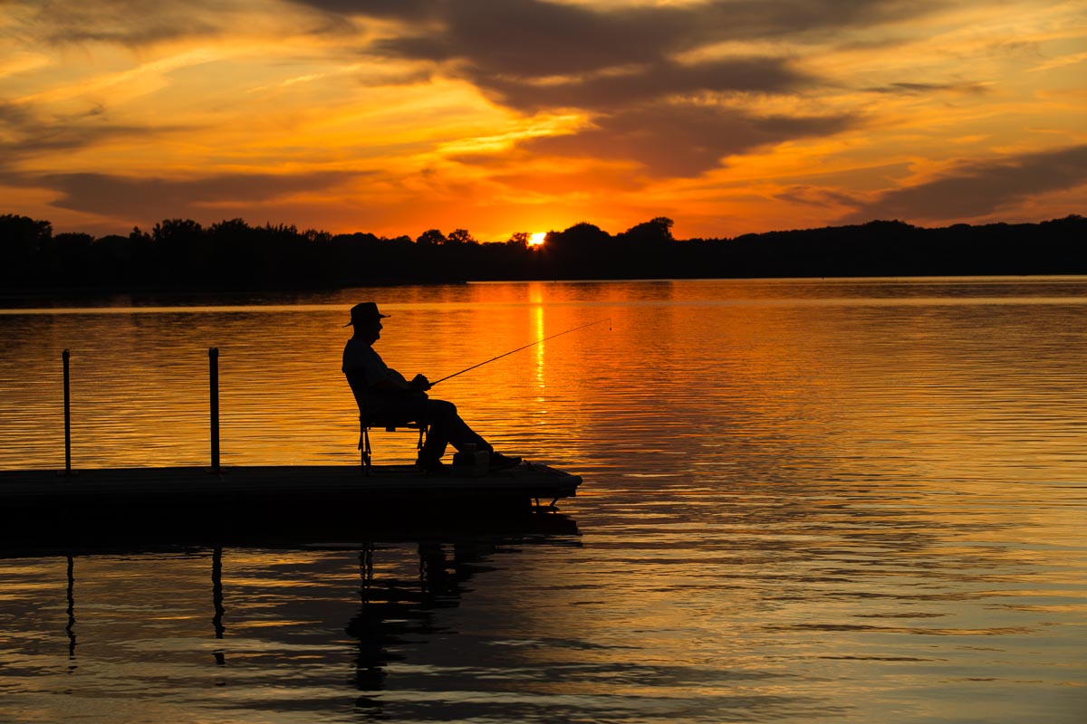 A fisherman sits on a dock, silhouetted against a sunset-lit lake at Willow Creek State Recreation Area near Pierce. Photo by Jenny Nguyen-Wheatley